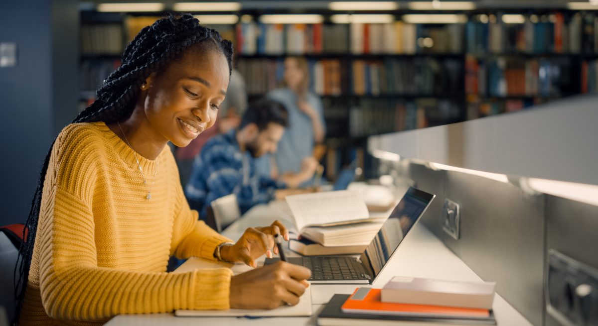 A young black woman in a yellow shirt researches with a laptop, pen and paper, in a university library.