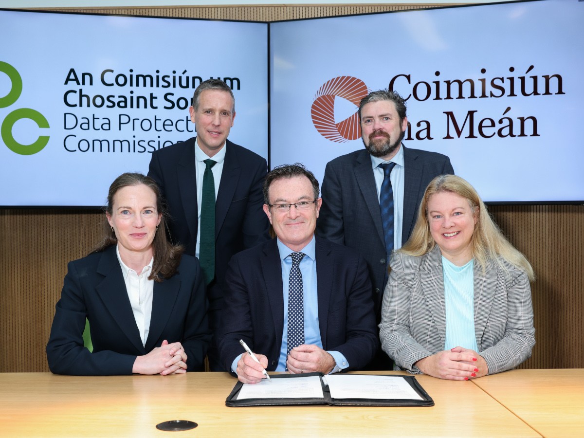 Two women and one man sit and two men stand behind them with an agreement paper on the table in front and the DPC and CnM logos on screens behind them.